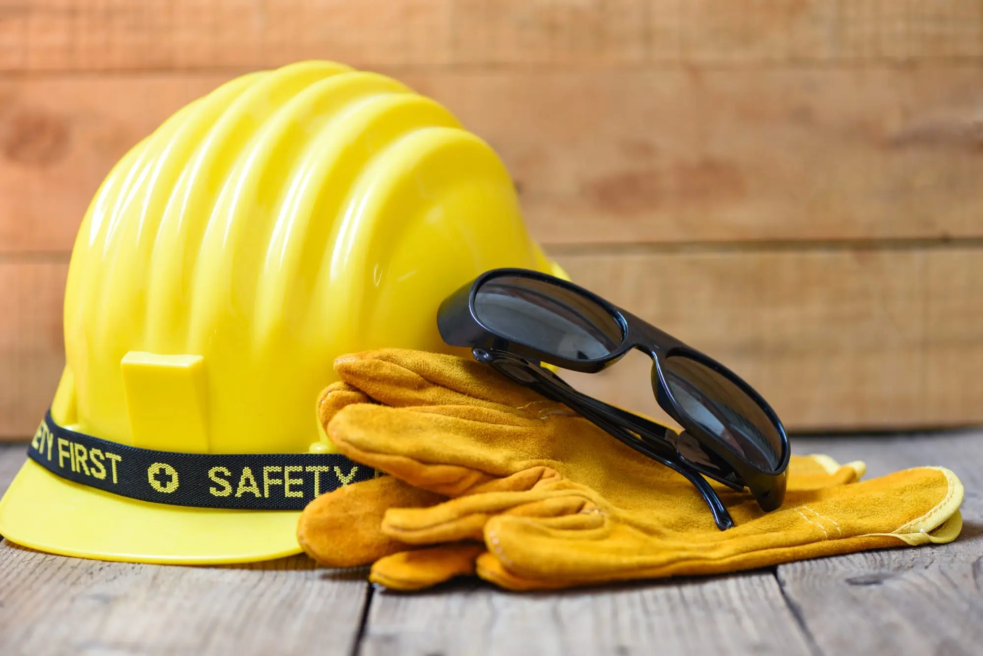 Casco de seguridad amarillo con la leyenda Safety First, guantes de cuero y gafas oscuras sobre una mesa de madera